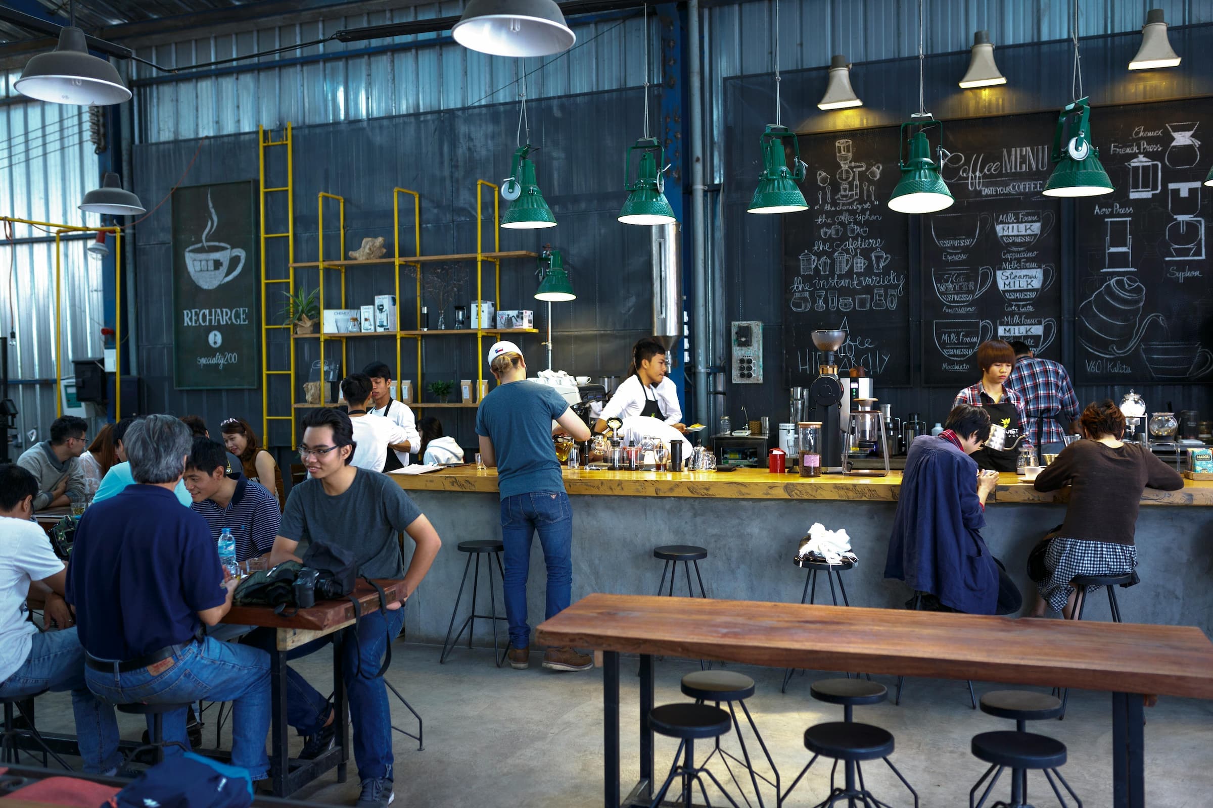 Baristas working behind a busy industrial-style coffee bar with customers seated and a chalkboard menu in the background